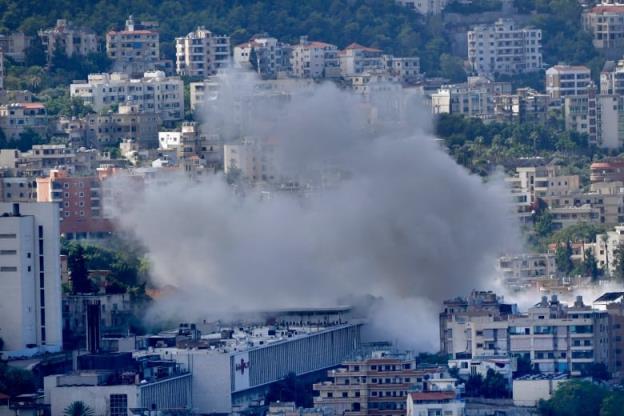 Multilevel buildings are shown in the hillside of a town, with large clouds of smoke seen rising in the air.